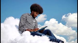 a 20 year old man with an afro is sitting on fluffy a cloud and reading a book in the middle of a bl