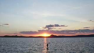 A young woman with long hair stands on a board triumphantly lifting up an oar at sunset.