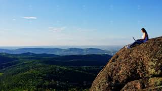 A female freelancer works sitting on the edge of a rocky cliff in the forest.